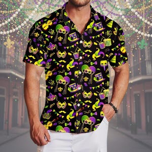 Pode incluir: Camisa preta de manga curta com um padrão vibrante com tema de Mardi Gras. O design apresenta máscaras, instrumentos musicais e símbolos festivos em amarelo, verde e roxo. A camisa é feita de tecido leve.