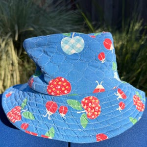 May include: A blue quilted bucket hat featuring a repeating pattern of red berries, green leaves, and light blue gingham apples. The fabric has a stitched diamond texture throughout the crown and brim, creating a soft, padded appearance.