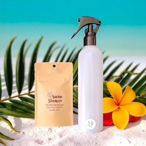 Puede incluir: Una botella de spray blanca con un gatillo negro, un pequeño paquete marrón con el texto "Swim Shower" y una flor amarilla y roja sobre una superficie arenosa. El fondo presenta hojas de palma y un cielo azul.