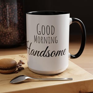 Puede incluir: Una taza de cerámica blanca con interior y asa negros. La taza tiene las palabras "GOOD MORNING Handsome" impresas en negro. Una cuchara, un macaron y granos de café están sobre una superficie de madera.