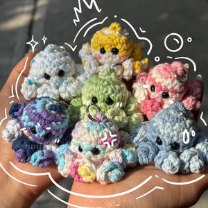 May include: A collection of handmade crochet octopus keychains in various colors, including white, yellow, green, pink, blue, and purple. Each octopus has a metal keyring attached and features black button eyes. The keychains are held in a person's hand.