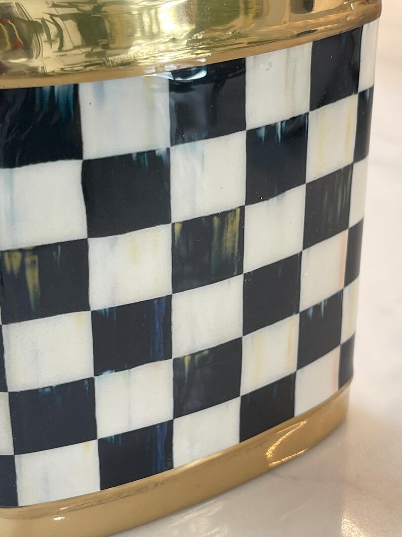 May include: A decorative container with a black and white checkered pattern. The top and base are trimmed with a gold-colored metal. The container has a glossy finish and a rounded rectangular shape.
