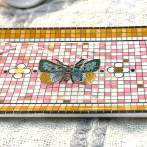 Puede incluir: Bandeja decorativa rectangular con dise&ntilde;o de mosaico. La bandeja presenta un patr&oacute;n de cuadros rosas y blancos con una mariposa colorida en el centro. El borde es un mosaico de azulejos amarillos y naranjas. Tambi&eacute;n hay flores decorativas.