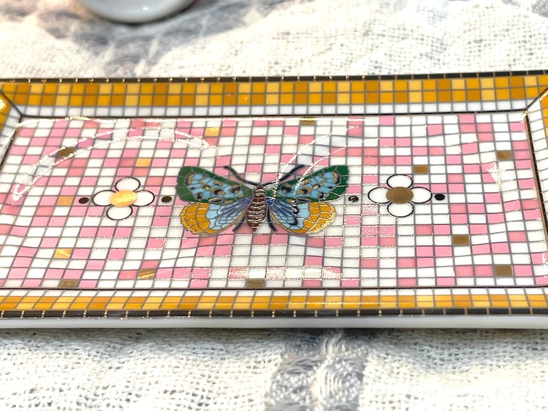 Puede incluir: Bandeja decorativa rectangular con dise&ntilde;o de mosaico. La bandeja presenta un patr&oacute;n de cuadros rosas y blancos con una mariposa colorida en el centro. El borde es un mosaico de azulejos amarillos y naranjas. Tambi&eacute;n hay flores decorativas.