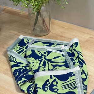 May include: Folded green and navy blue kitchen towel with a floral pattern. The towel has a white border with a gray stripe. The towel is on a wooden surface.