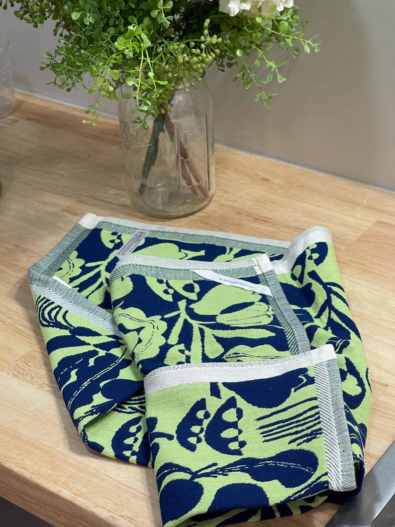 May include: Folded green and navy blue kitchen towel with a floral pattern. The towel has a white border with a gray stripe. The towel is on a wooden surface.