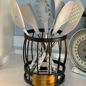 Può includere: Un portautensili cilindrico in metallo nero con una targhetta color ottone recante la scritta MacKenzie-Childs, contenente cinque utensili da cucina in silicone bianco con manici a scacchi, tra cui una schiumarola, una spatola, un mestolo e una paletta.