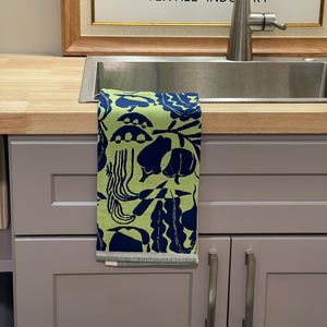 May include: A green and navy blue patterned kitchen towel hangs over a stainless steel sink. The towel features a repeating design of abstract shapes. The background includes a framed sign with text about arts and crafts.