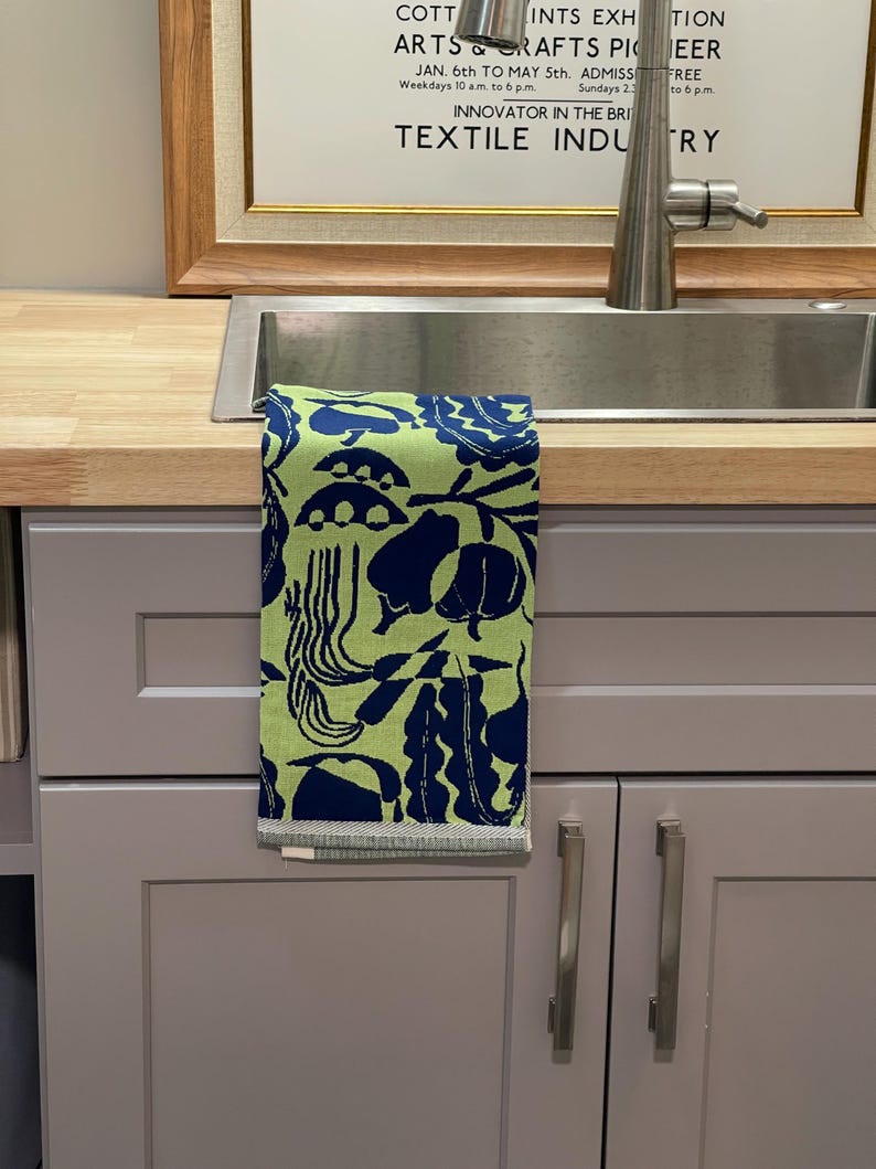 May include: A green and navy blue patterned kitchen towel hangs over a stainless steel sink. The towel features a repeating design of abstract shapes. The background includes a framed sign with text about arts and crafts.