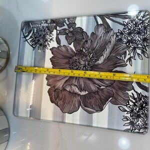 Puede incluir: Bandeja rectangular de vidrio con dise&ntilde;o floral. La bandeja presenta una flor grande y detallada en tonos grises y marrones, sobre un fondo a rayas. La bandeja mide aproximadamente 28 cm de largo.
