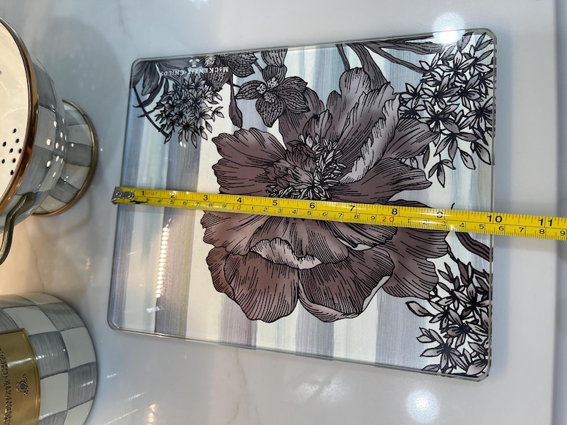 Puede incluir: Bandeja rectangular de vidrio con dise&ntilde;o floral. La bandeja presenta una flor grande y detallada en tonos grises y marrones, sobre un fondo a rayas. La bandeja mide aproximadamente 28 cm de largo.