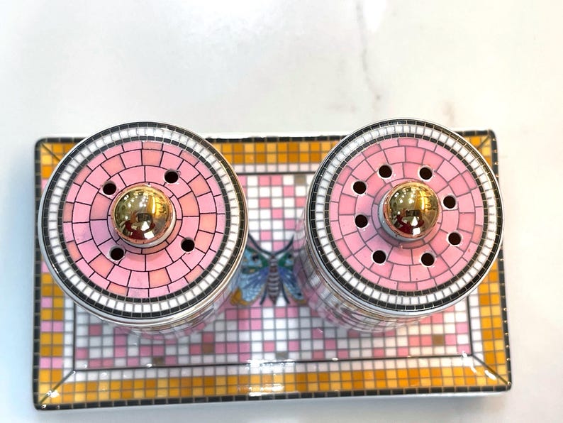 Puede incluir: Un juego decorativo de salero y pimentero con tapas de mosaico rosa y blanco y detalles dorados. El juego incluye dos saleros y una bandeja rectangular con un dise&ntilde;o de mariposa.
