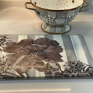Puede incluir: Tabla de cortar rectangular de vidrio con dise&ntilde;o floral. La tabla presenta una flor grande y detallada en tonos marr&oacute;n y gris, sobre un fondo a rayas. Acentos florales decorativos enmarcan los bordes. Un colador con borde dorado en el fondo.