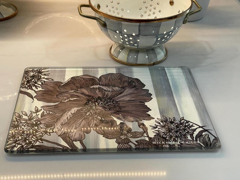 Puede incluir: Tabla de cortar rectangular de vidrio con dise&ntilde;o floral. La tabla presenta una flor grande y detallada en tonos marr&oacute;n y gris, sobre un fondo a rayas. Acentos florales decorativos enmarcan los bordes. Un colador con borde dorado en el fondo.
