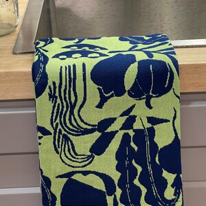 May include: A folded kitchen towel with a repeating pattern of navy blue botanical shapes on a light green background. The towel has a striped border at the bottom. The towel is folded over a stainless steel sink.