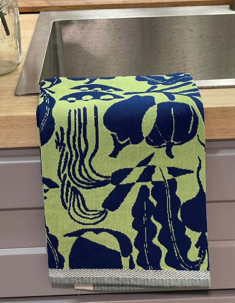 May include: A folded kitchen towel with a repeating pattern of navy blue botanical shapes on a light green background. The towel has a striped border at the bottom. The towel is folded over a stainless steel sink.