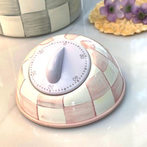 Könnte beinhalten: Ein rosa-weiß karierter Küchentimer mit hellviolettem Zifferblatt und rosa Sockel. Der Timer ist kuppelförmig und hat ein weißes Zifferblatt mit Zahlen. Im Hintergrund befindet sich ein passender karierter Behälter.