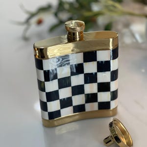 May include: A decorative flask with a gold-colored cap and trim. The body of the flask features a black and white checkered pattern. The flask's cap is unscrewed and placed next to the flask.