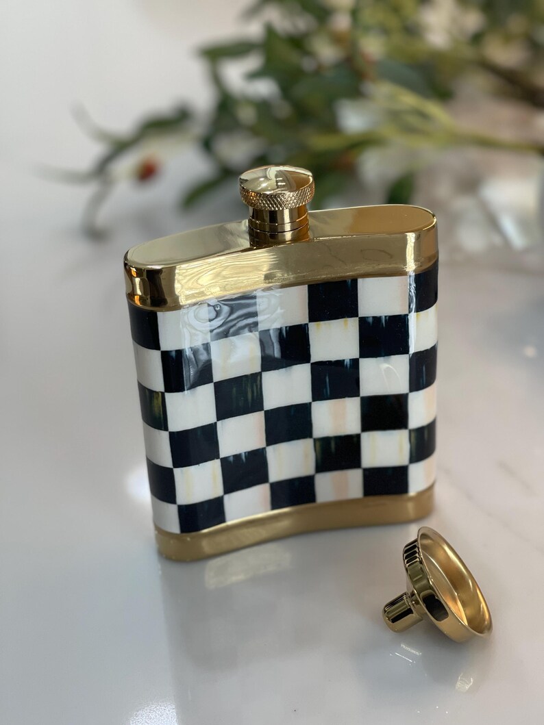 May include: A decorative flask with a gold-colored cap and trim. The body of the flask features a black and white checkered pattern. The flask's cap is unscrewed and placed next to the flask.