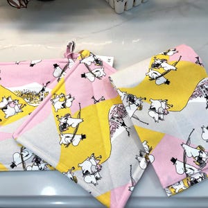 May include: Two quilted fabric pot holders featuring a repeating pattern of white Moomin characters on a background of pink, yellow, and light gray geometric shapes. The design includes small black top hats and detailed illustrations of a house.