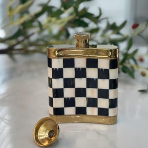 May include: A decorative flask with a black and white checkered pattern. The flask has a gold-colored rim and a gold-colored cap. A gold-colored funnel sits next to the flask. The flask is rectangular and appears to be made of ceramic.