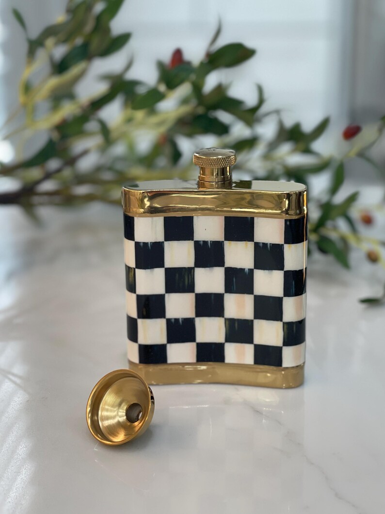 May include: A decorative flask with a black and white checkered pattern. The flask has a gold-colored rim and a gold-colored cap. A gold-colored funnel sits next to the flask. The flask is rectangular and appears to be made of ceramic.