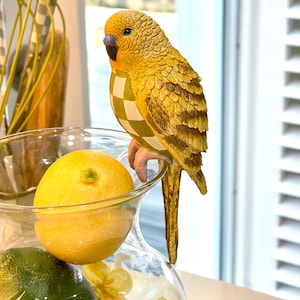 May include: A decorative yellow parakeet figurine with a checkered pattern on its chest, perched on a glass vase. The vase contains lemons and limes. The parakeet has detailed feather textures and a blue eye.