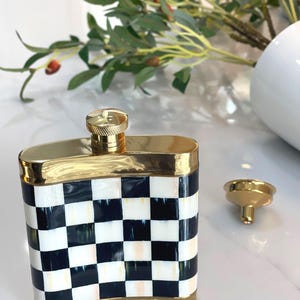 May include: A decorative flask with a gold-colored rim and cap. The flask features a black and white checkered pattern on the front and sides. A small gold funnel sits next to the flask.