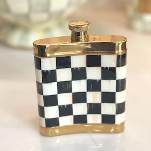May include: A decorative flask with a black and white checkered pattern. The flask has gold-colored trim around the top, bottom, and cap. The cap is also gold-colored. The flask is rectangular in shape.
