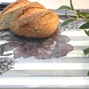Puede incluir: Una hogaza de pan crujiente, cortada para revelar el interior, descansa sobre una bandeja decorativa. El pan es de color marr&oacute;n dorado con hierbas visibles. La bandeja presenta un dise&ntilde;o floral con rayas grises y blancas.