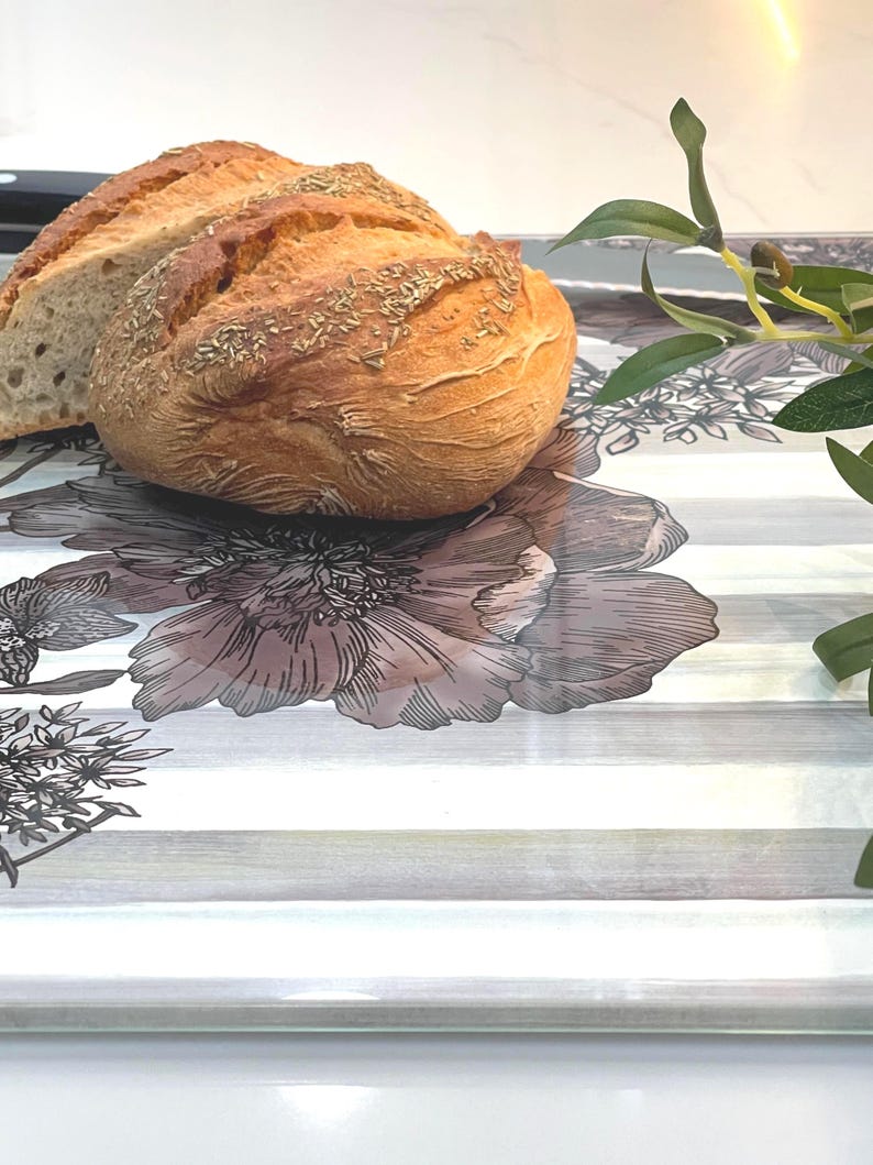 Puede incluir: Una hogaza de pan crujiente, cortada para revelar el interior, descansa sobre una bandeja decorativa. El pan es de color marr&oacute;n dorado con hierbas visibles. La bandeja presenta un dise&ntilde;o floral con rayas grises y blancas.