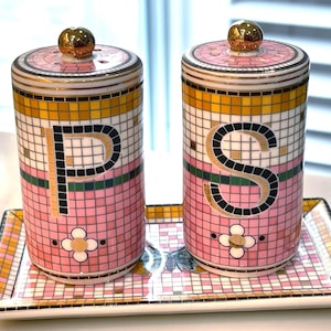 Puede incluir: Un juego de salero y pimentero rosa y dorado con una bandeja a juego. Los saleros son cil&iacute;ndricos con tapas doradas y presentan las letras "P" y "S". El juego tiene un dise&ntilde;o de mosaico con un motivo floral.