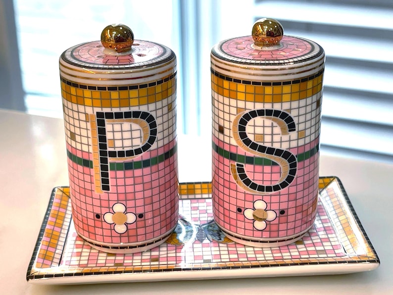 Puede incluir: Un juego de salero y pimentero rosa y dorado con una bandeja a juego. Los saleros son cil&iacute;ndricos con tapas doradas y presentan las letras "P" y "S". El juego tiene un dise&ntilde;o de mosaico con un motivo floral.