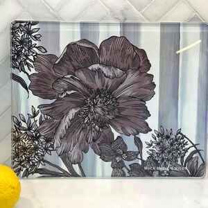 Puede incluir: Tabla de cortar rectangular de vidrio con dise&ntilde;o floral. La tabla presenta una flor grande y detallada en tonos grises y morados, con peque&ntilde;os acentos florales. El fondo tiene rayas verticales grises y blancas.