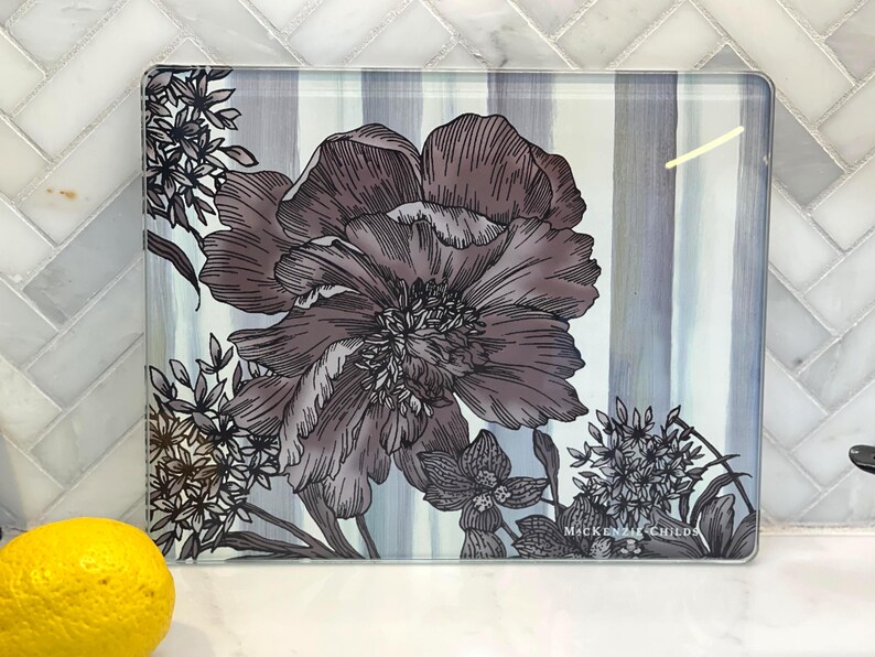 Puede incluir: Tabla de cortar rectangular de vidrio con dise&ntilde;o floral. La tabla presenta una flor grande y detallada en tonos grises y morados, con peque&ntilde;os acentos florales. El fondo tiene rayas verticales grises y blancas.