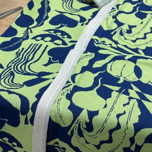 May include: Close-up of a fabric with a repeating pattern of blue and green organic shapes. A white woven trim runs along the edge. The fabric is likely used for home decor or apparel.