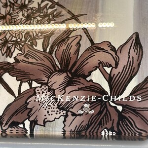 Puede incluir: Funda de tel&eacute;fono decorativa con un dise&ntilde;o floral en tonos marr&oacute;n y blanco. La funda tiene las palabras "MacKenzie-Childs" impresas, junto con un peque&ntilde;o emblema. El dise&ntilde;o incluye ilustraciones detalladas de flores y follaje.