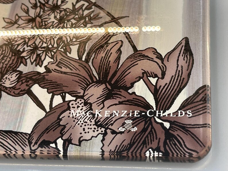 Puede incluir: Funda de tel&eacute;fono decorativa con un dise&ntilde;o floral en tonos marr&oacute;n y blanco. La funda tiene las palabras "MacKenzie-Childs" impresas, junto con un peque&ntilde;o emblema. El dise&ntilde;o incluye ilustraciones detalladas de flores y follaje.