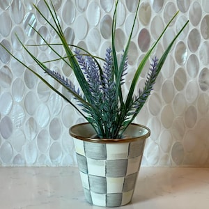 Puede incluir: Una maceta de metal pequeña y cónica con un patrón de cuadros grises y blancos pintado a mano y un borde dorado metálico. La maceta contiene un arreglo decorativo de hojas de hierba verde artificial y tallos florales de color violeta.