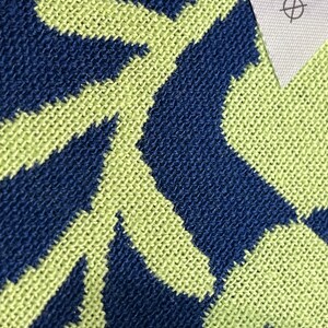 May include: Close-up of a knitted textile with a repeating geometric pattern in navy blue and light green. A white tag with text and symbols is visible in the upper right corner.