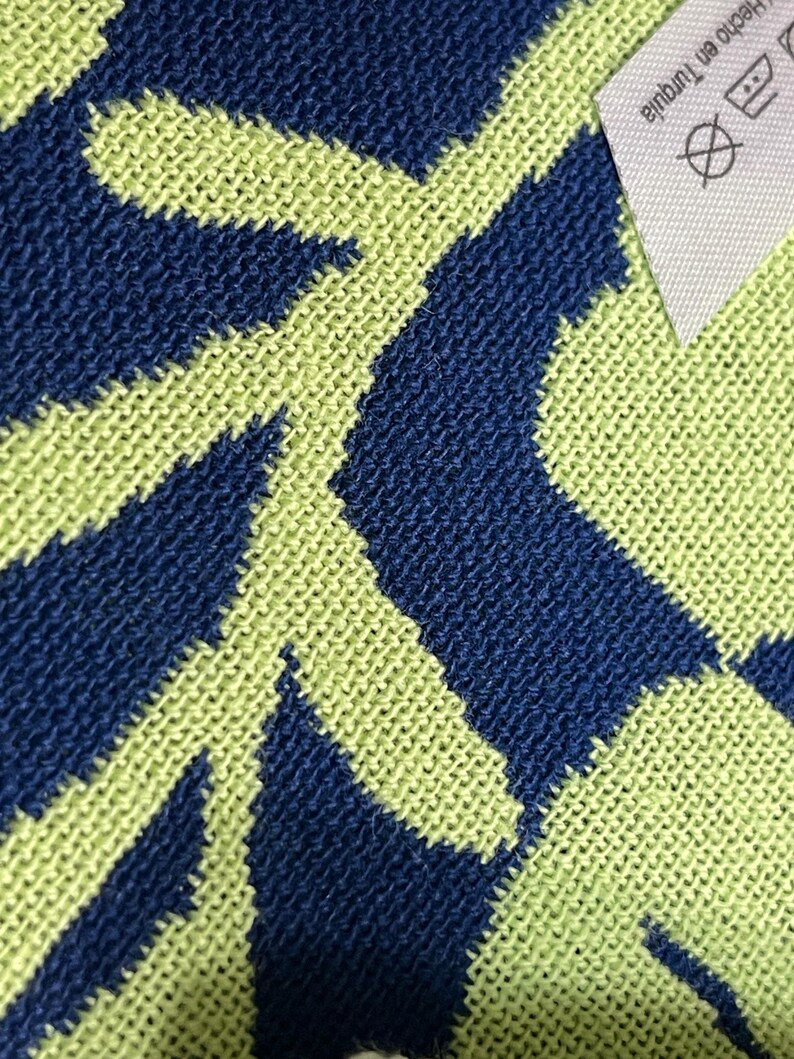 May include: Close-up of a knitted textile with a repeating geometric pattern in navy blue and light green. A white tag with text and symbols is visible in the upper right corner.