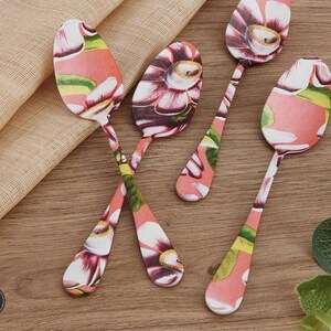 May include: Set of floral-patterned spoons. Each spoon features a pink, white, and green floral design on the spoon bowl and handle. The spoons are arranged on a light brown surface, creating a visually appealing arrangement.