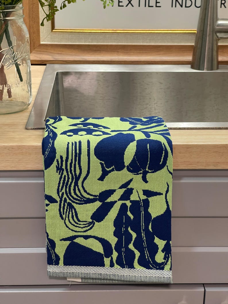 May include: A folded kitchen towel with a vibrant pattern of navy blue botanical shapes on a light green background. The towel is made of a soft fabric and has a striped border.