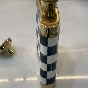 May include: A decorative flask with a gold-colored cap and trim. The body of the flask features a black and white checkered pattern. The flask is made of metal and has a screw-on cap. The cap is detached in the image.