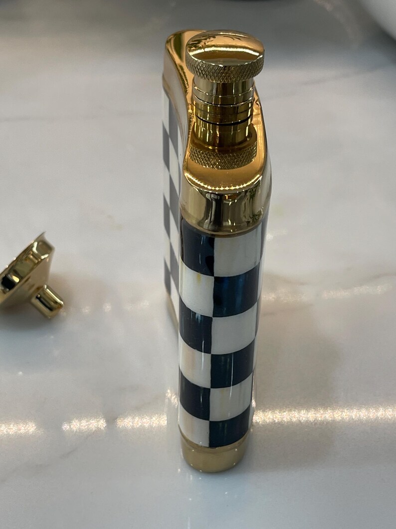 May include: A decorative flask with a gold-colored cap and trim. The body of the flask features a black and white checkered pattern. The flask is made of metal and has a screw-on cap. The cap is detached in the image.