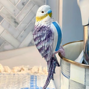 May include: A decorative parakeet figurine with a white head, yellow accents, and a blue beak. The bird has a blue stripe on its side and purple and white feathered wings. It is perched on the edge of a checkered container.