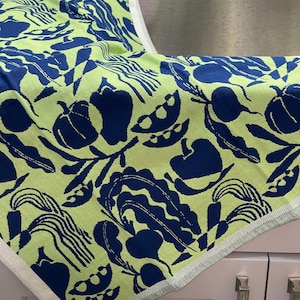 May include: A folded kitchen towel with a vibrant pattern of navy blue vegetables and foliage on a light green background. The towel has a white and light green border and is made of a soft, absorbent material.