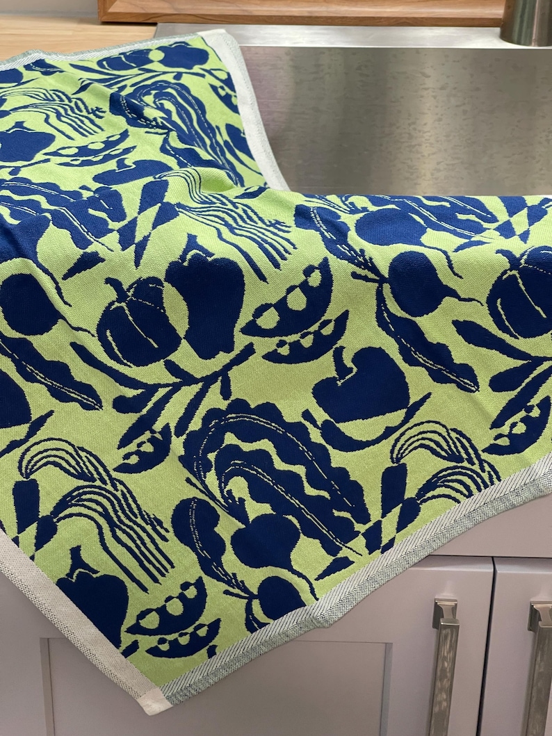 May include: A folded kitchen towel with a vibrant pattern of navy blue vegetables and foliage on a light green background. The towel has a white and light green border and is made of a soft, absorbent material.