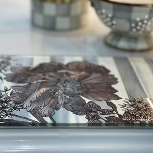 Puede incluir: Bandeja rectangular de vidrio con dise&ntilde;o floral. La bandeja presenta una flor grande y detallada en tonos grises y marrones, sobre un fondo a rayas. Los bordes son transparentes y la bandeja tiene un acabado brillante.