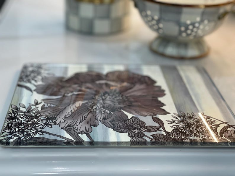 Puede incluir: Bandeja rectangular de vidrio con dise&ntilde;o floral. La bandeja presenta una flor grande y detallada en tonos grises y marrones, sobre un fondo a rayas. Los bordes son transparentes y la bandeja tiene un acabado brillante.